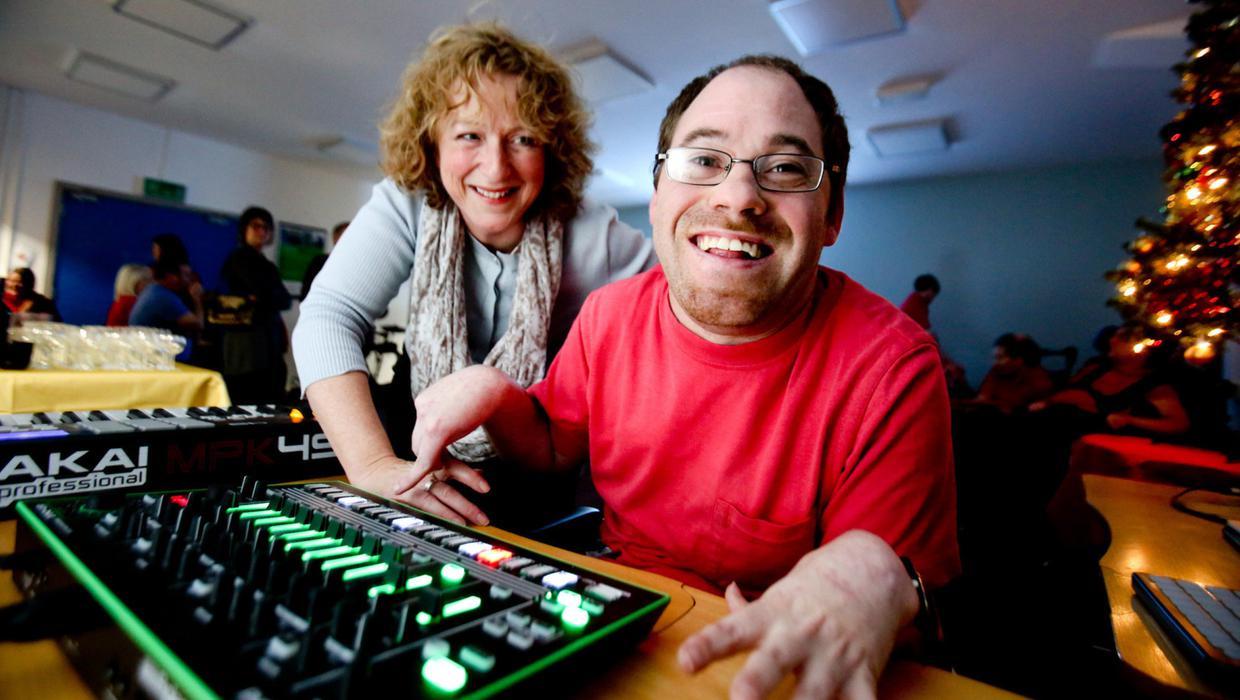 Musician Tim smiles at the camera, while playing Roland TR-8, accompanied by his carer, who smiles at him.