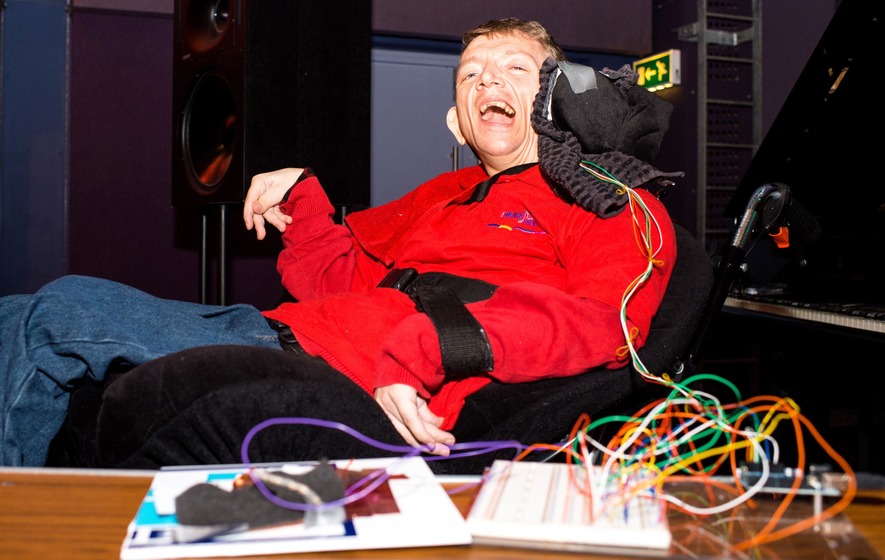 Musician Ray Hamilton looks and smiles at the camera. He has various wires connecting to his wheelchair to an interface at the desk in front of him.