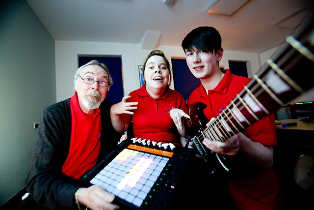 Thee musicians smile at the camera. They hold an Ableton Push and an electric guitar. They all wear the same looking shirt.