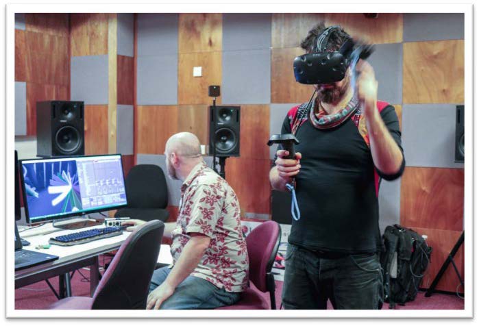 Participant wearing VR gogle and joysticks, tests newly built accessible virtual instrument, while standing up. The tutor and designer Damian Mills is sitting in front of a computer, where you see the musician's point of view. Indoor, studio at SARC - Queen's University, Belfast.
