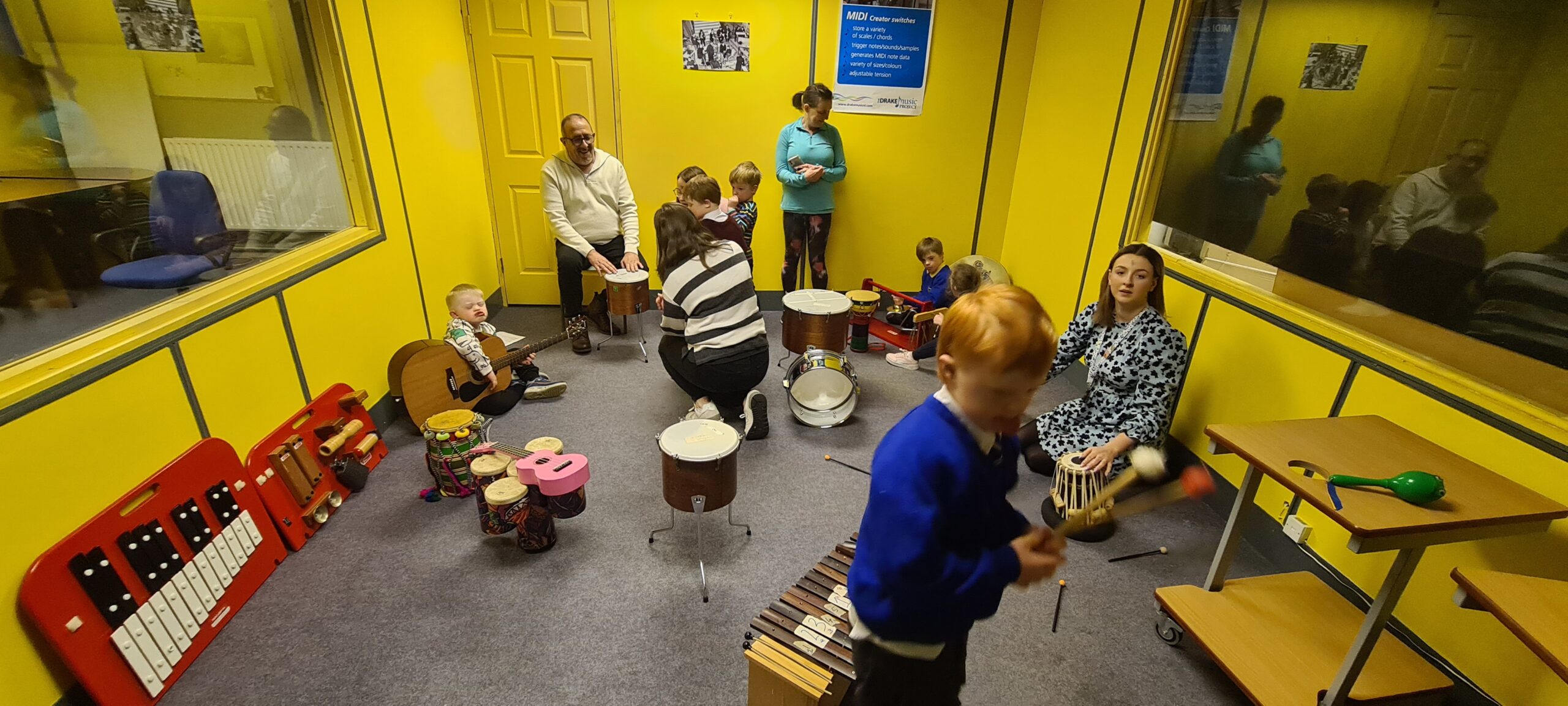 Tutor Nikos, siting down, plays a small drum with a group of 6 children, who also play different intruments, such as guitar and percussion intruments. They accompained by 3 ladies who also assist the children. during a the Down Right music session at Drake's studio in Newry.
