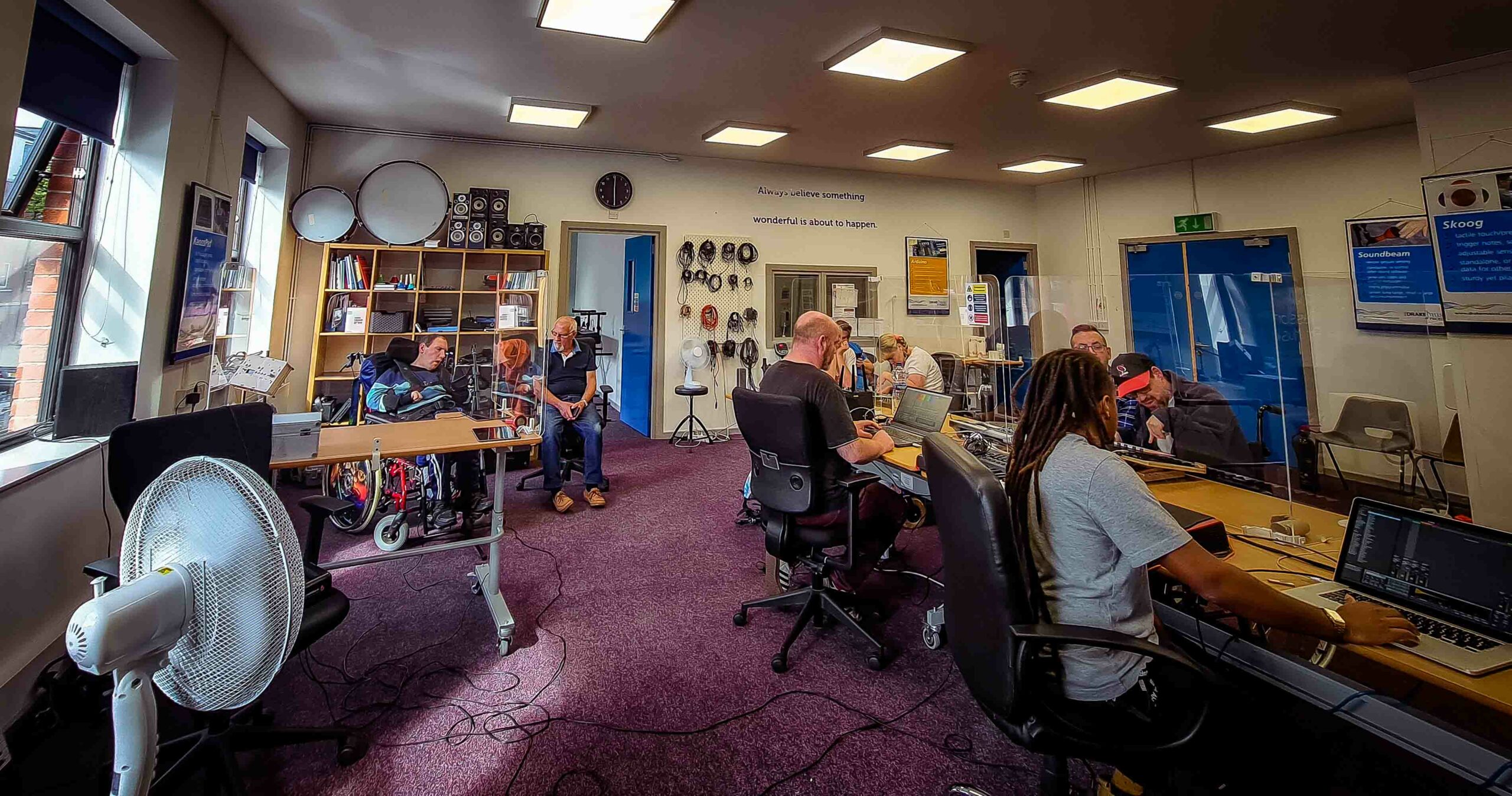 Wide shot of musicians Eoin Fitzpatrick, Marylouise McCord and Tim Leathem play electronic instruments during a Wired Ensemble Session, accompanied by their family assinting members and the tutors Henrique Franco and Damian Mills, at Drake's Belfast Studio